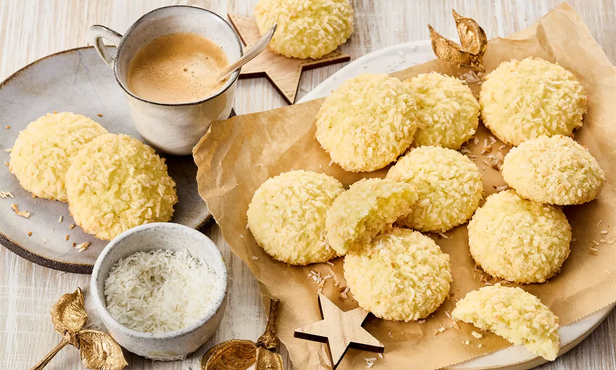 Kokos-Cookies: goldbraune, lockere Kekse mit weißen Kokosraspeln, auf einem Holzteller gestapelt und mit zusätzlichem Kokos bestreut