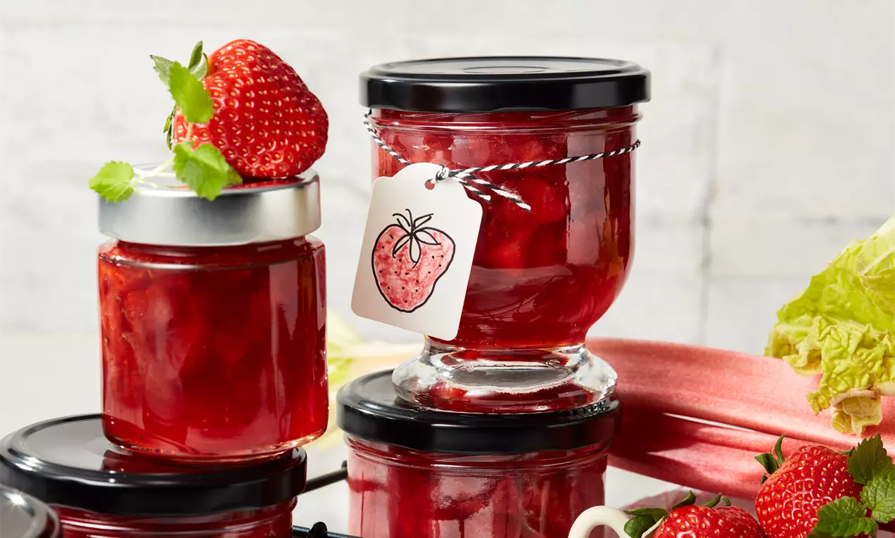 Erdbeer-Rhabarber-Konfitüre in klaren Gläsern mit glänzend rotem Inhalt, dekoriert mit frischen Erdbeeren und grünen Blättern