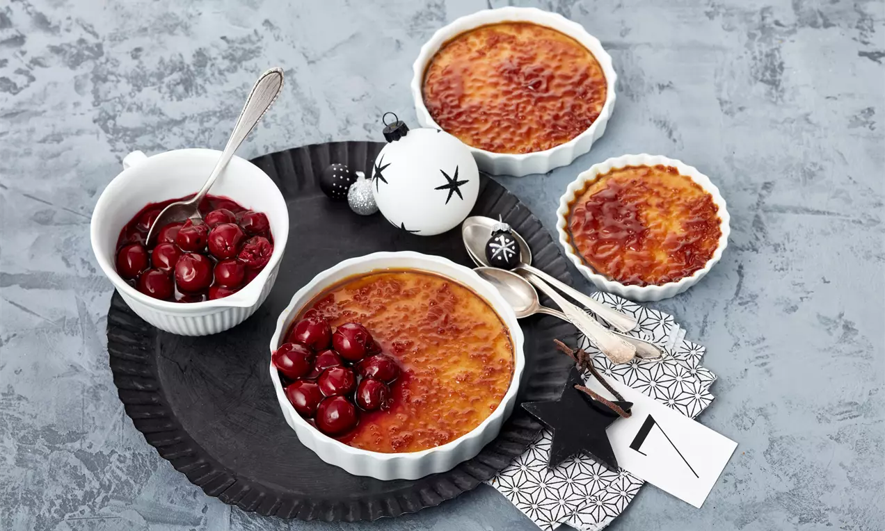 Zimt-Crème-Brûlée mit karamellisierter Oberfläche in weißen Förmchen, serviert mit glänzenden Glühweinkirschen auf schwarzem Teller