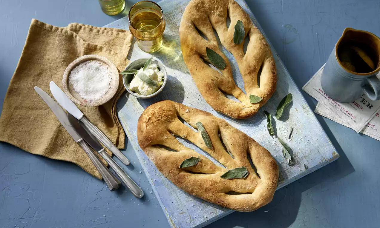 Zwei goldbraune, blattförmige Salbei-Zitronen-Fougasse mit eingesetzten Salbeiblättern auf blauem Brett, flankiert von Beilagen und Besteck