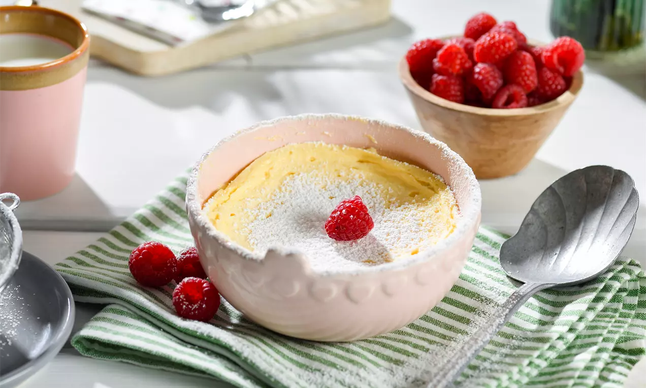 Goldgelber Käsekuchen mit weißem Puderzucker, serviert in einer rosa Schale, garniert mit einer Himbeere auf gestreiftem Stofftuch