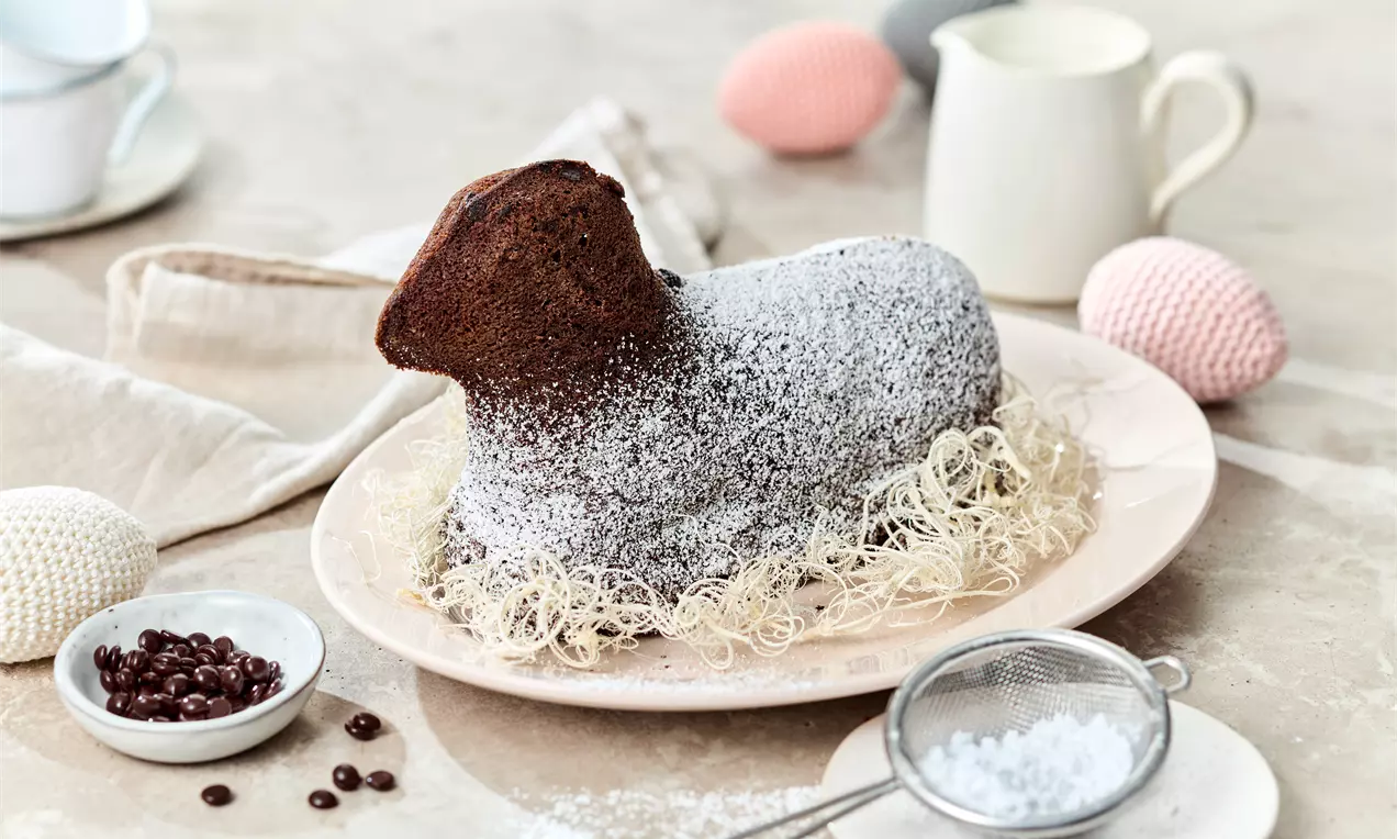 Schokoladiges Lamm in dunkler, leicht rauer Teigtextur, mit Puderzucker bestäubt, auf Ostergras auf einem Teller arrangiert