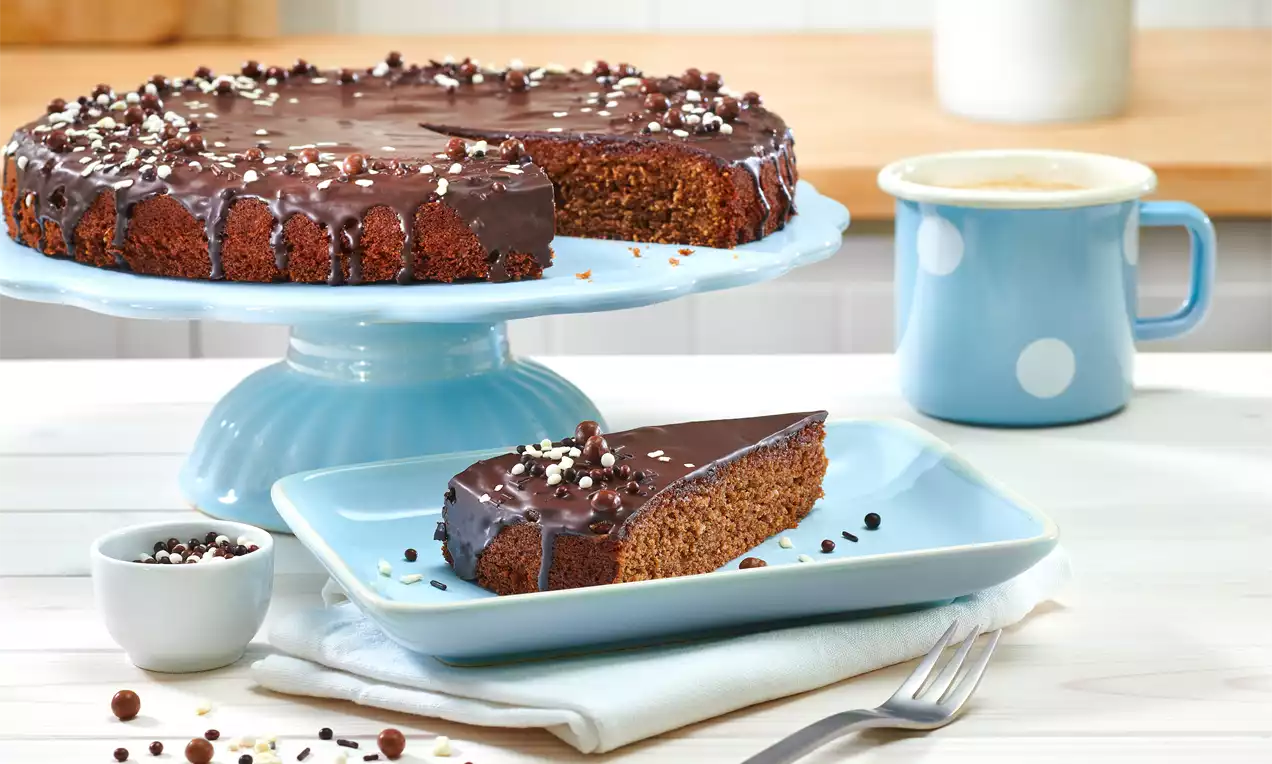 Nougatkuchen mit glänzender Schokoglasur und Schokoladenstreuseln auf blauer Tortenplatte, ein Stück auf einem Teller mit Gabel angerichtet