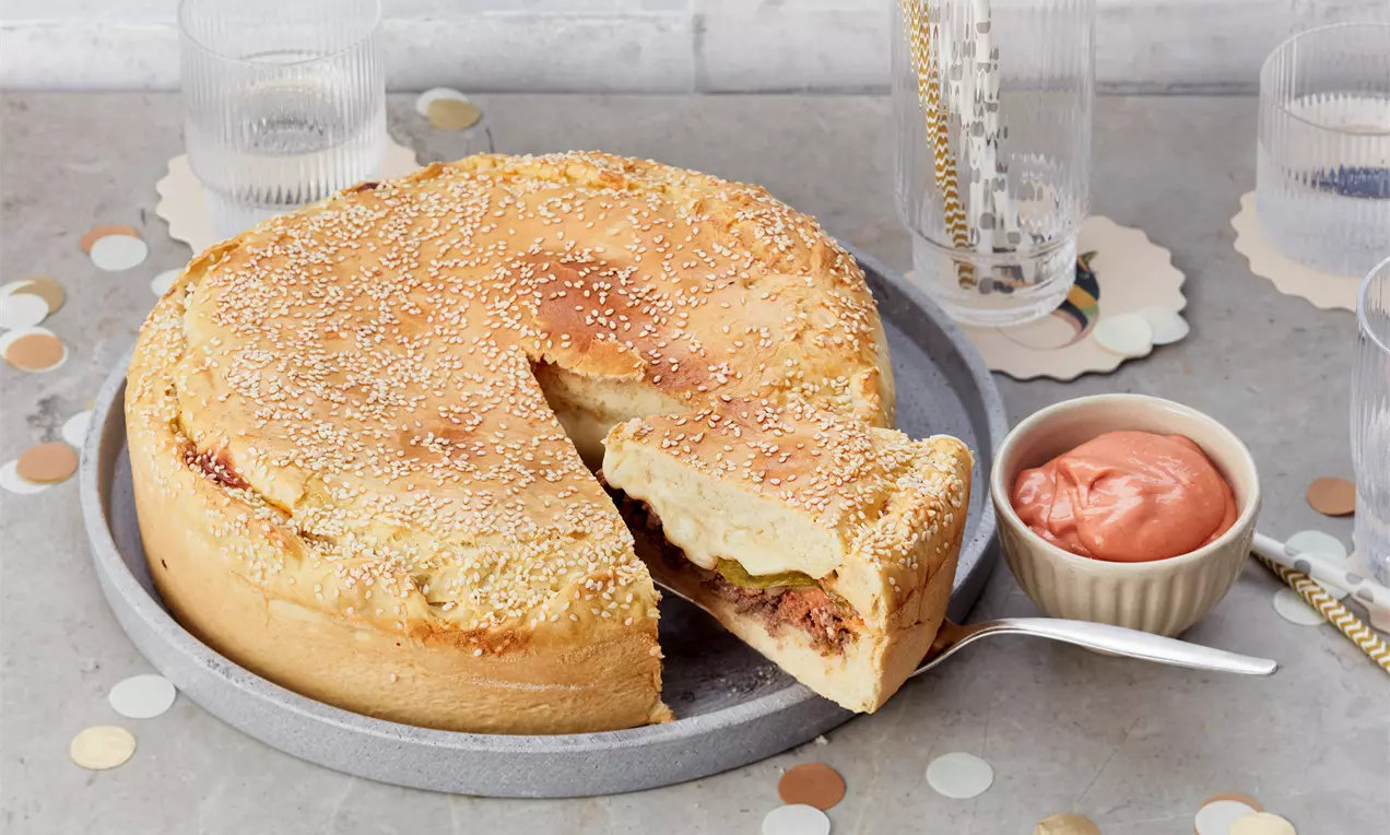 Runder Cheeseburger-Kuchen mit goldbrauner Sesamkruste, angeschnitten auf grauem Teller, serviert mit rosafarbener Sauce in einem kleinen Schälchen
