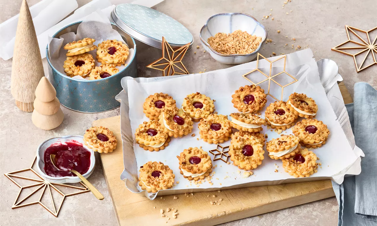 Goldbraune Frankfurter-Kranz-Plätzchen mit rotem Marmeladenkern und knusprigen Krokantstücken auf weißem Backpapier auf einem Holzbrett