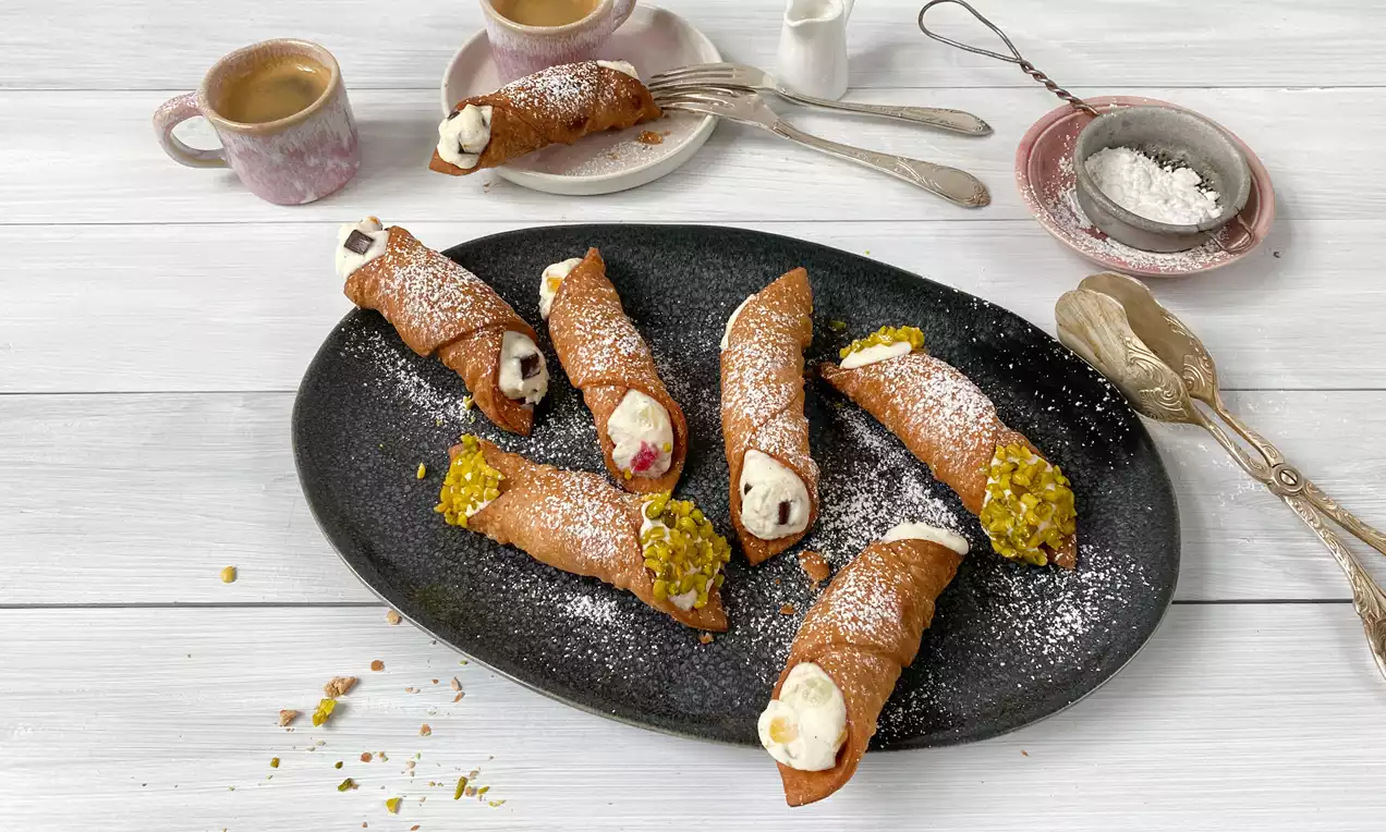 Cannoli mit goldbraun-knusprigem Teig, gefüllt mit weißer Creme, teils mit Pistazien und Puderzucker garniert, auf dunkler Platte serviert