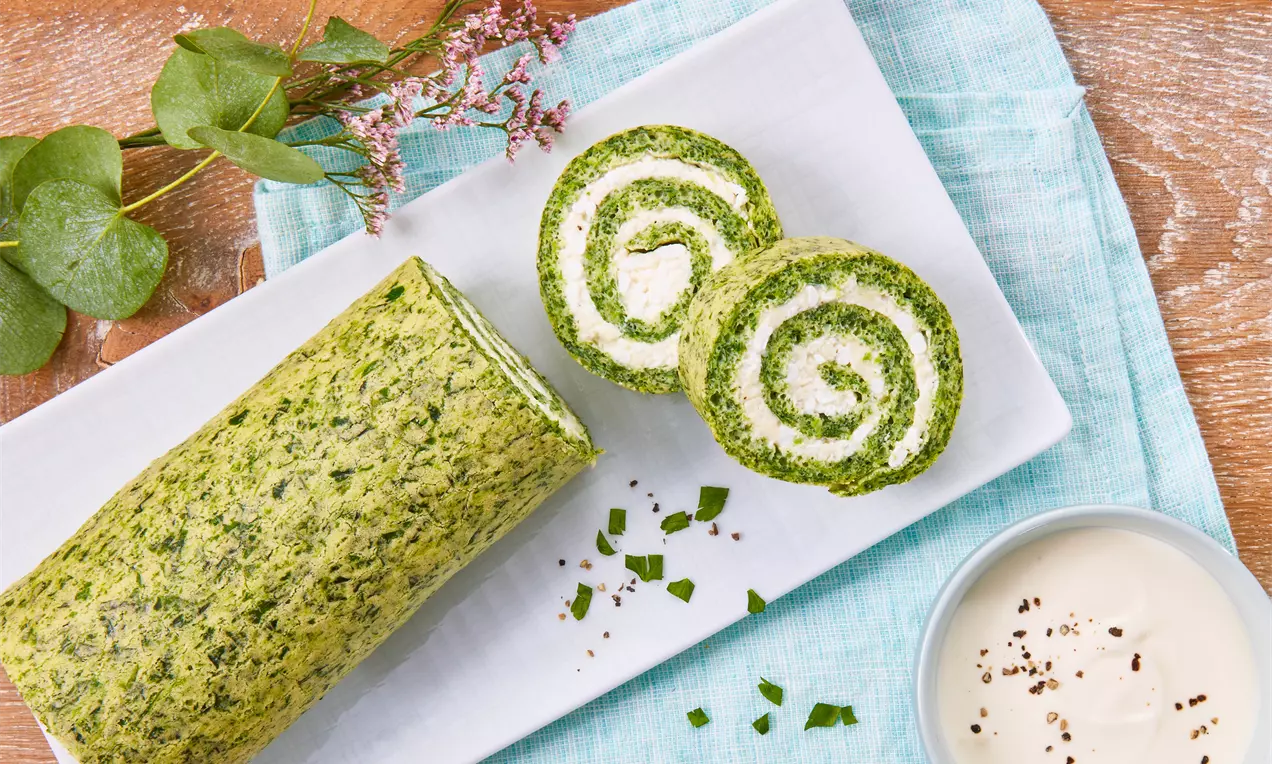 Spinatrolle mit grünlich-marmorierter Oberfläche und Frischkäse-Füllung in Spiralmuster, angerichtet auf weißer Platte mit Kräuterdekor