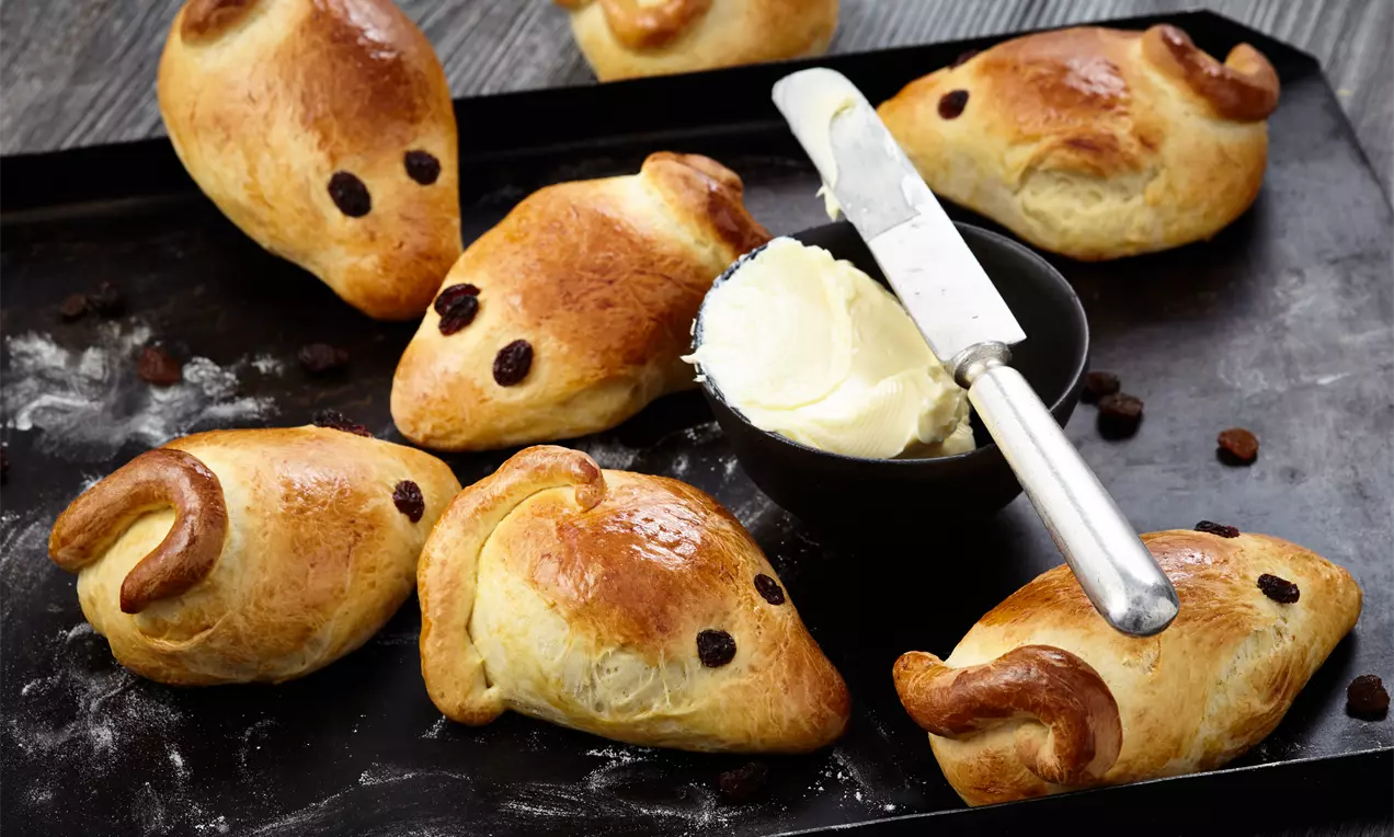 Goldbraune Hefemäuse mit rosinenartigen Augen und geschwungenen Schwänzen auf dunklem Blech neben einer Schale mit cremiger Butter