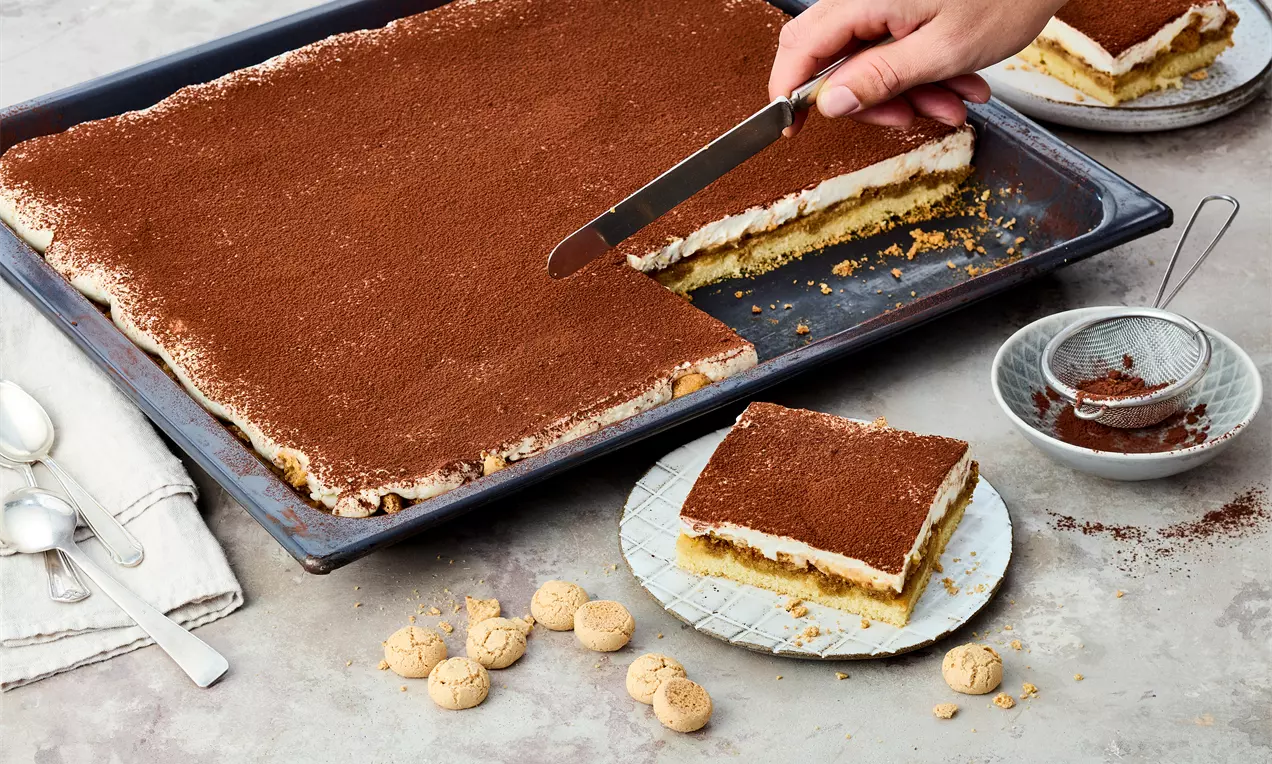 Ein rechteckiger Tiramisu Kuchen auf einem Backblech, mit saftigem Boden, getränkt in Espresso-Amaretto, belegt mit knusprigen Amarettini und einer dicken Schicht cremiger Mascarpone-Quark-Creme