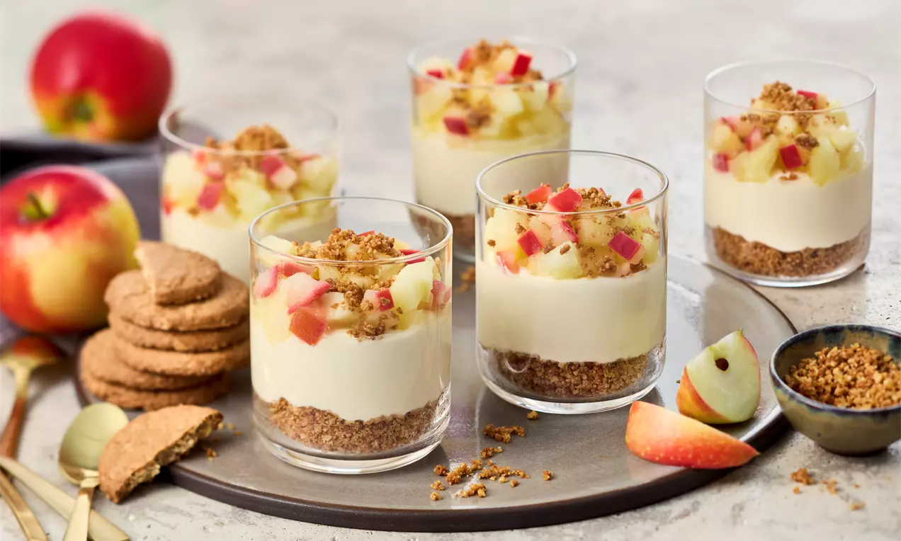 Apfel-Käsekuchen-Dessert im Glas, geschichtet mit Keksboden, cremiger Füllung und Apfelstückchen, garniert mit Streuseln. Mehrere Gläser auf einem Teller, daneben Äpfel, Kekse und eine Schale Streusel