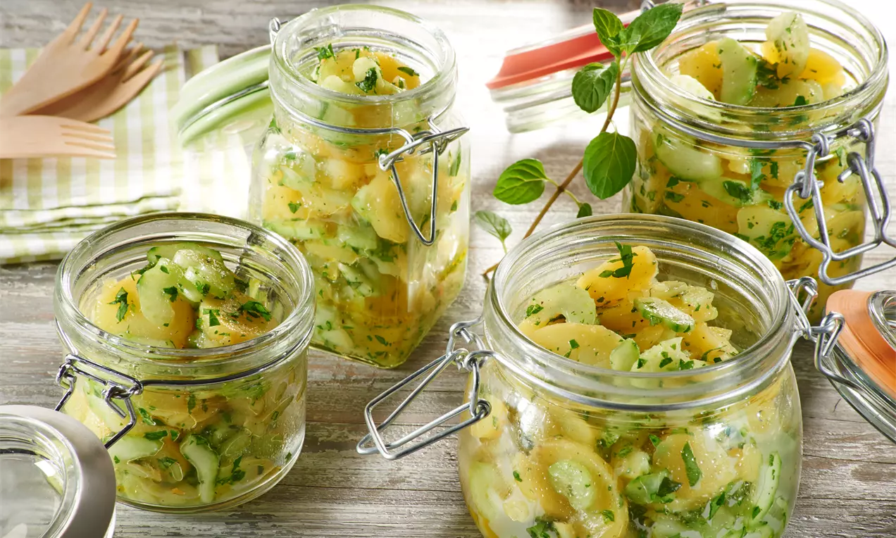 Goldgelber Kartoffelsalat mit grüner Petersilie, Gurkenscheiben und Zwiebelringen in vier offenen Einmachgläsern mit Bügelverschluss drapiert