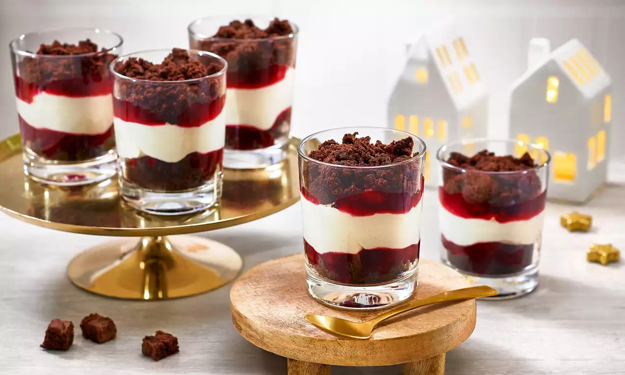Schichtdessert in klaren Gläsern mit roter Fruchtsoße, weißer Creme und braunen Kuchenstücken auf Holz und goldener Platte arrangiert