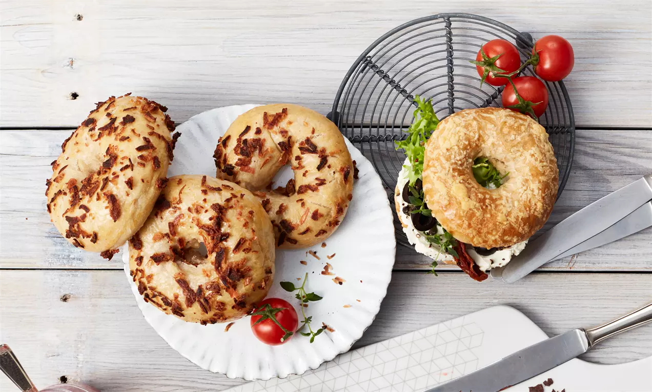 Bagels mit knusprigen Belägen und unterschiedlicher Textur auf Tellern und Schneidebrettern, dekoriert mit Kirschtomaten und Dip-Schalen