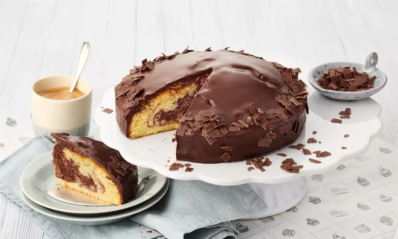 Marmorierter Kuchen mit glänzender Schokoladenglasur und Schokoraspeln, eine Scheibe angeschnitten, serviert auf weißer Tortenplatte mit Kaffee und Untertasse