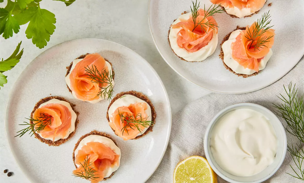 Kleine Canapés mit braunem Boden, weißer Creme, zartrosa Lachsrosen und frischem Dill auf weißen Tellern arrangiert