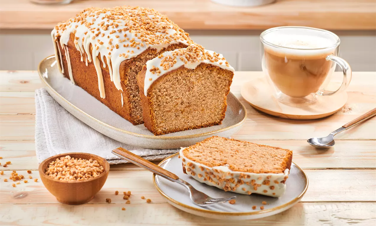 Goldbrauner Zimtkuchen mit heller Glasur und Krokant dekoriert, serviert auf einem Teller mit Cappuccino und Krokantschale daneben
