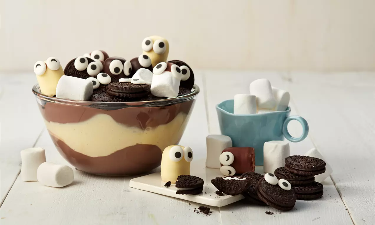 Ein Schicht-Dessert mit hellgelber Vanille- und brauner Schokocreme in einer Glasschale, verziert mit Marshmallows, Keksen und Zuckeraugen
