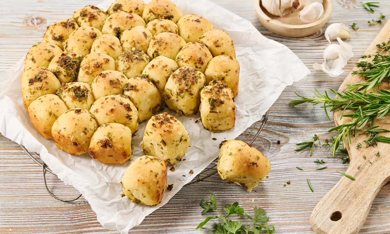 Runde, goldbraune Knoblauchbrot-Bällchen mit Kräutern bestreut, auf Backpapier drapiert, umgeben von frischen Kräutern und Knoblauchknollen