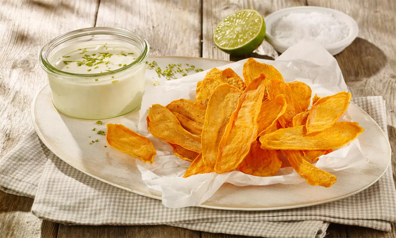 Knusprig goldene Süßkartoffel-Chips auf weißem Pergamentpapier mit cremigem Limetten-Dip im Glas, dekoriert mit Limettenzesten