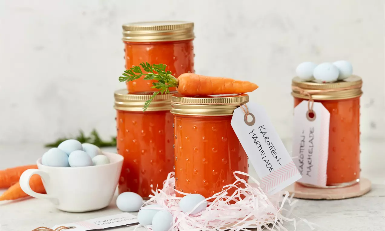 Gläser mit leuchtend orangefarbener Karottenmarmelade, goldenen Deckeln, Etiketten, dekoriert mit Karotten und blauen Zuckereiern