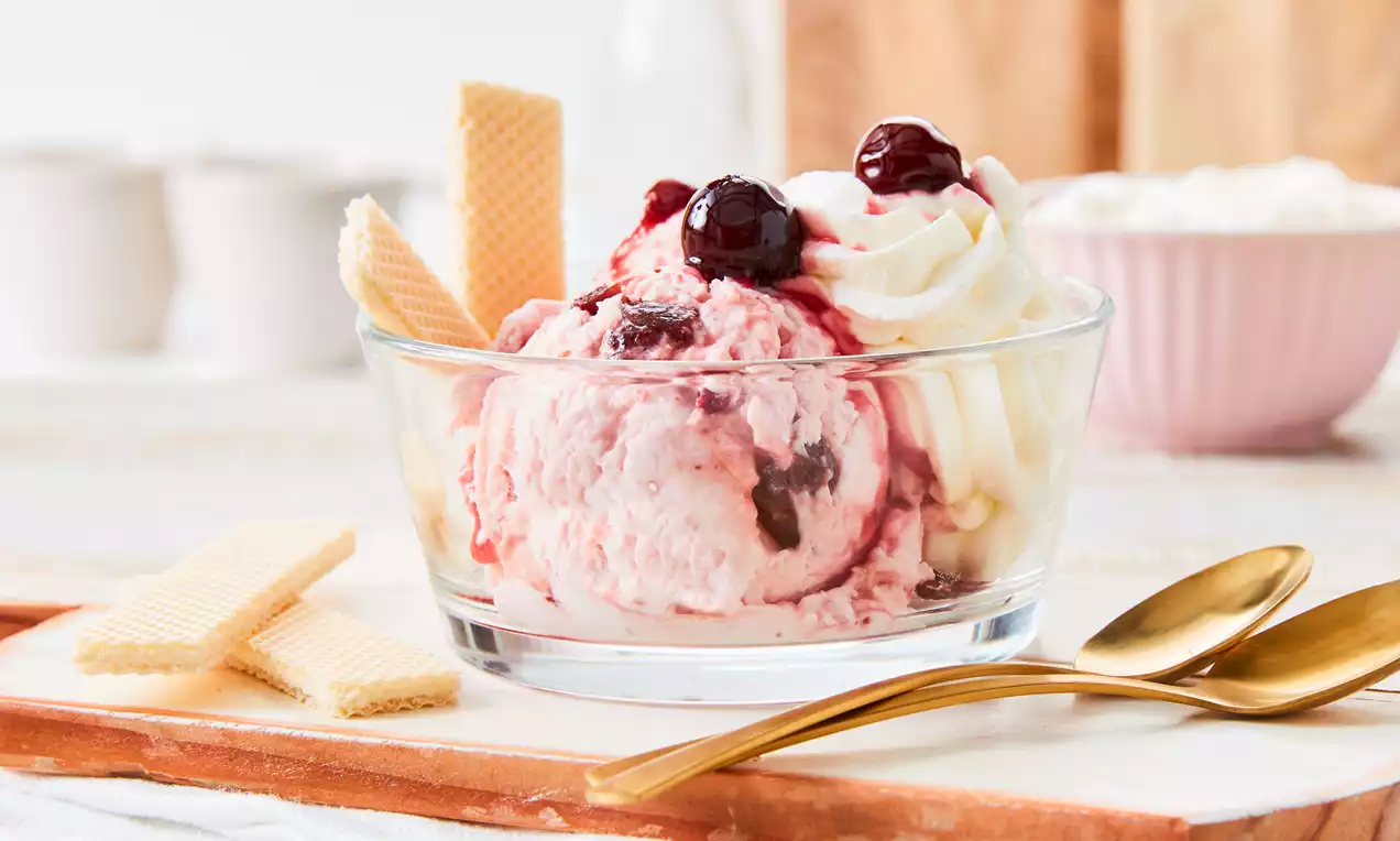 Ein Glasbecher mit rosa Amarena-Eis, dunklen Kirschen, weißer Sahne, roter Kirschsoße, goldenen Löffeln und Waffeln auf hellem Holzbrett