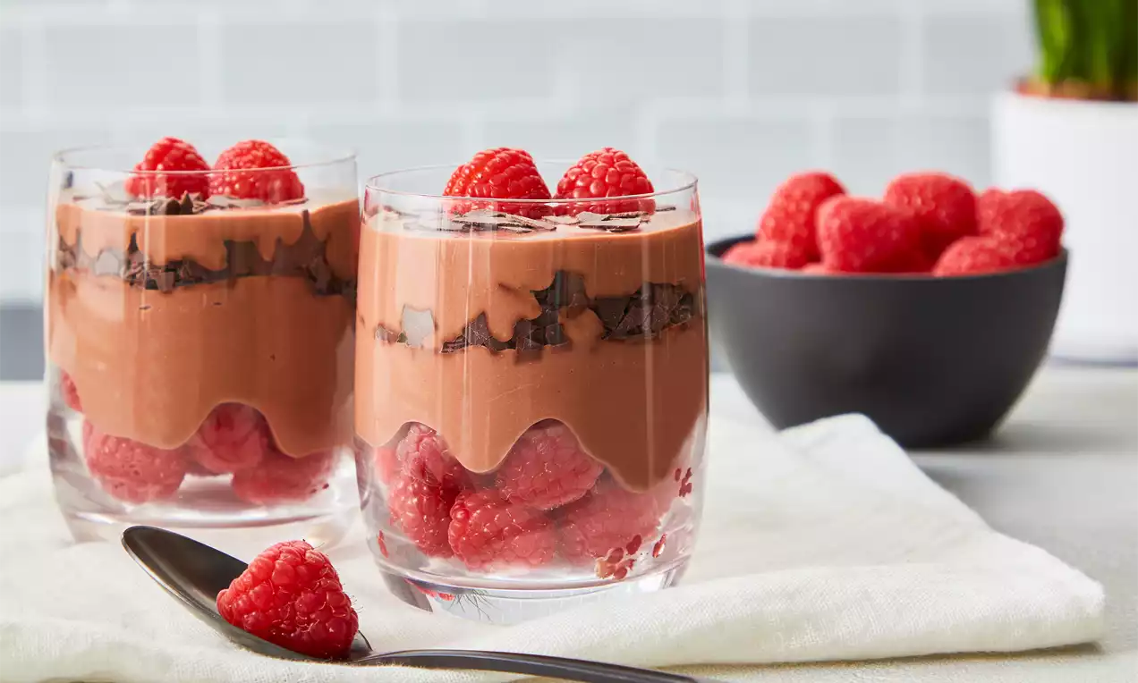 Zweischichtige Mascarpone-Schokocreme in Gläsern mit frischen Himbeeren und Schokoraspeln, dekoriert mit Himbeeren auf einem weißen Tuch