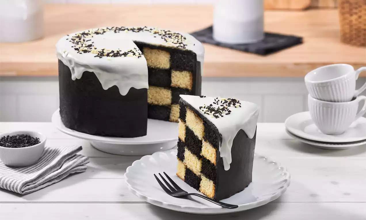 Schachbrett-Torte mit schwarzem Fondant, weißer Glasur und Schokostreuseln, angeschnitten auf einem Teller neben Tassen und Beilagen