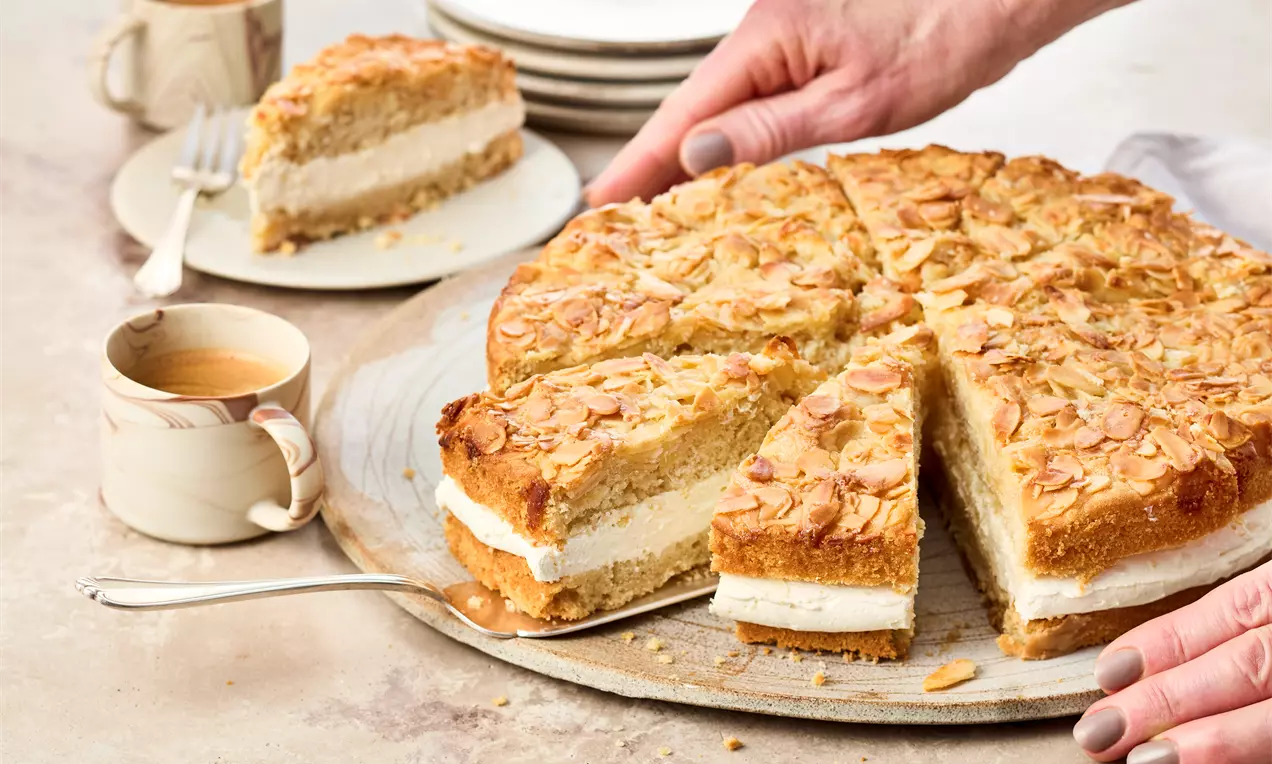 Veganer Bienenstich auf einer runden Platte, goldbraune Oberfläche mit Mandelblättchen, helle Cremefüllung sichtbar. Ein Stück ist herausgeschnitten, daneben Teller, Gabeln und eine Tasse Kaffee.