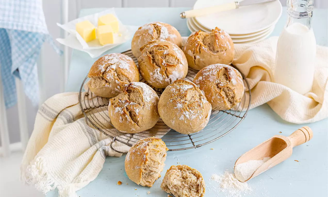 Goldbraune Sauerteig-Brötchen mit rissiger Kruste liegen auf einem runden Gitter, umgeben von Butterwürfeln, Mehl und Milchflasche