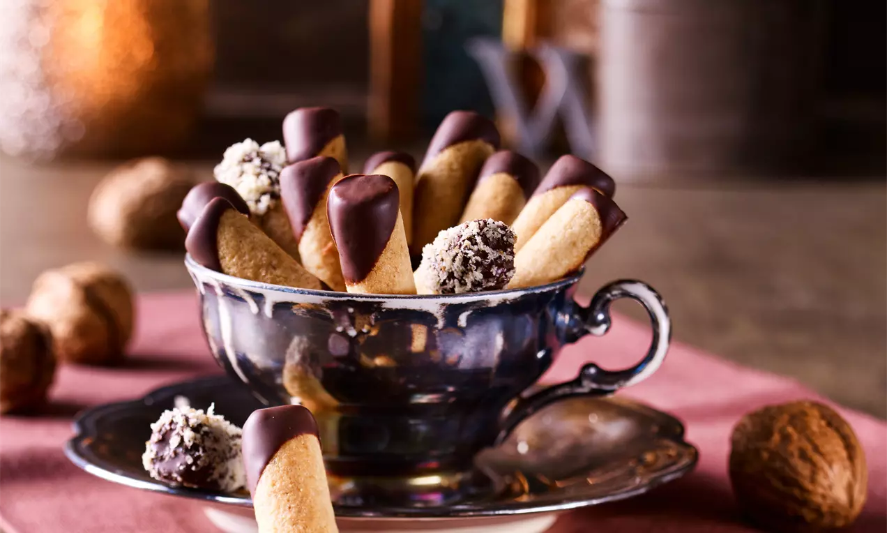 Walnuss-Makronen-Stäbchen mit dunkler Schokoladenspitze in einer glänzenden Tasse auf rosa Stoff, umgeben von Walnüssen und Trüffeln