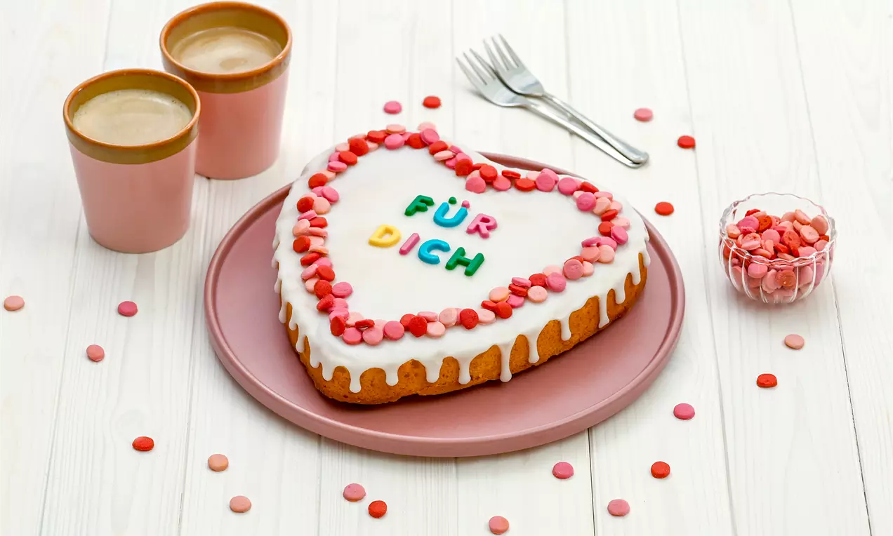 Herzförmiger Rührkuchen mit weißem Zuckerguss, bunter Verzierung und der Aufschrift „Für Dich“ auf einem rosa Teller mit Deko und Bechern