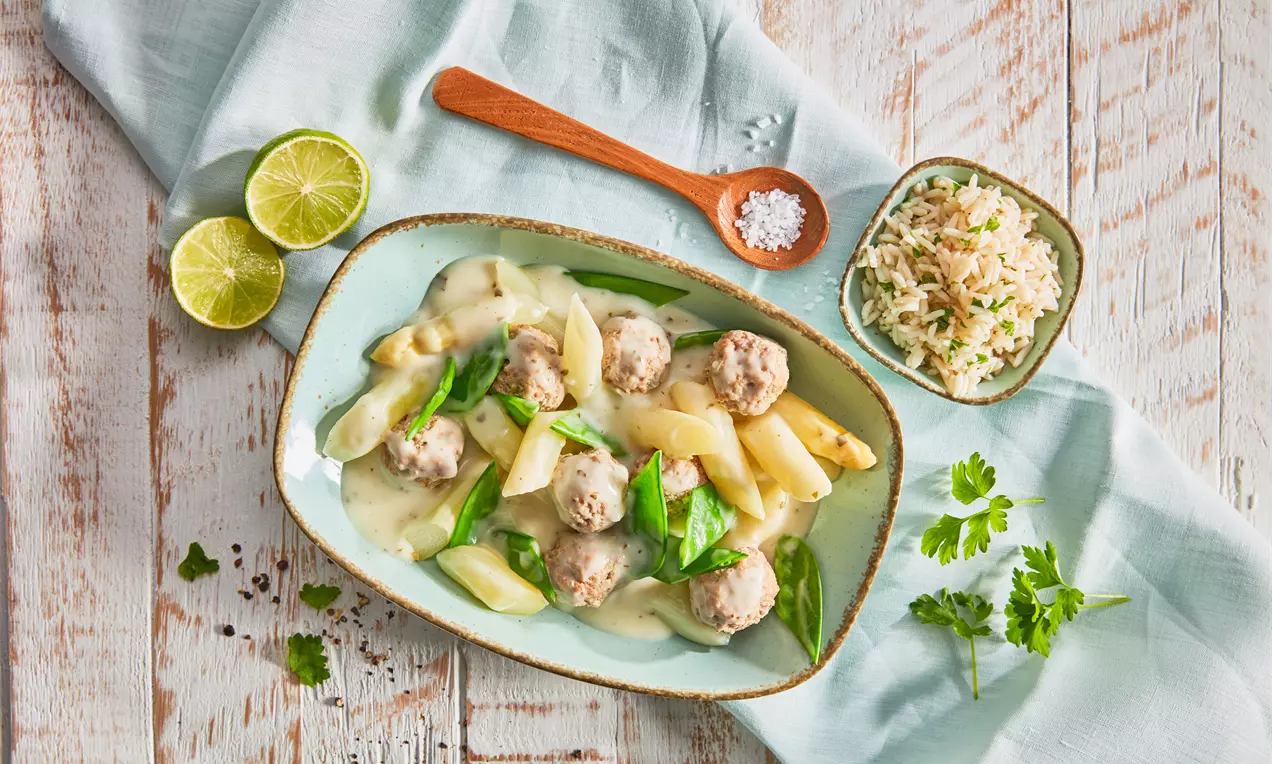 Spargelragout mit zarten Hackfleischbällchen in heller cremiger Sauce, garniert mit frischen grünen Blättern auf einem länglichen Teller