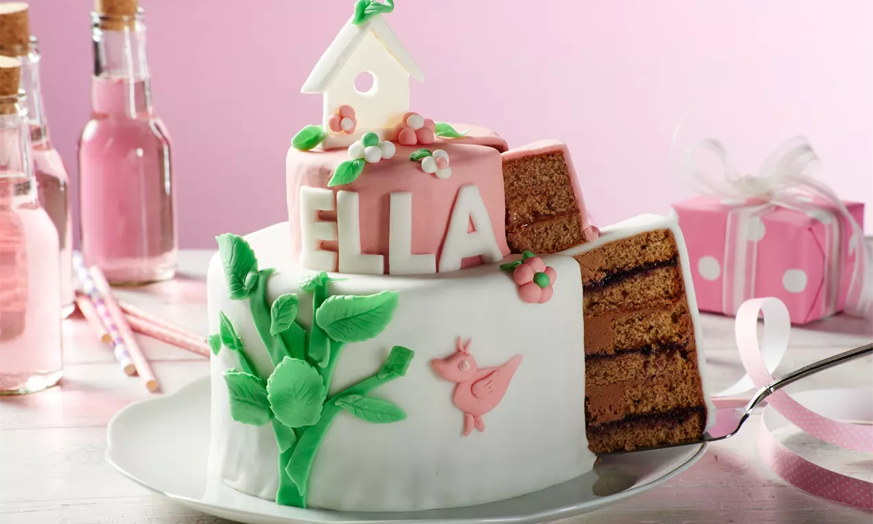 Zweistöckige Torte mit weißem Fondant, rosa Dekoren, blättrigem Baum und Vogel, angeschnitten zeigt sie braune Schichten mit Füllung