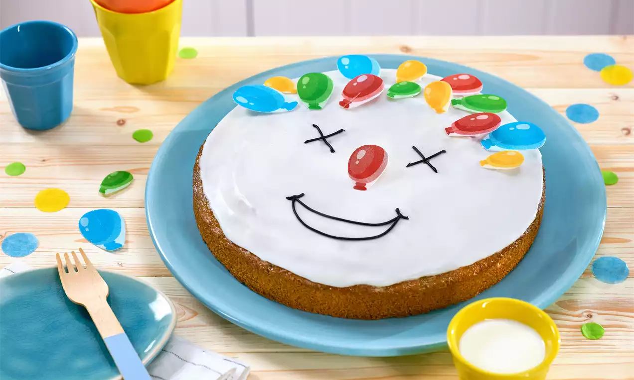 Runder Kuchen mit weißer Zuckerglasur, dekoriert mit bunten Luftballon-Zuckerstücken, schwarzen Linien als Gesicht auf blauem Teller
