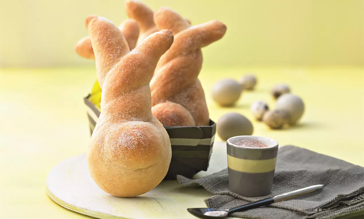 Goldbraune Dinkel-Häschen mit weicher Oberfläche und gebogenen Ohren, arrangiert in einem Streifenkorb auf hellem Untergrund, daneben Eier und Espresso