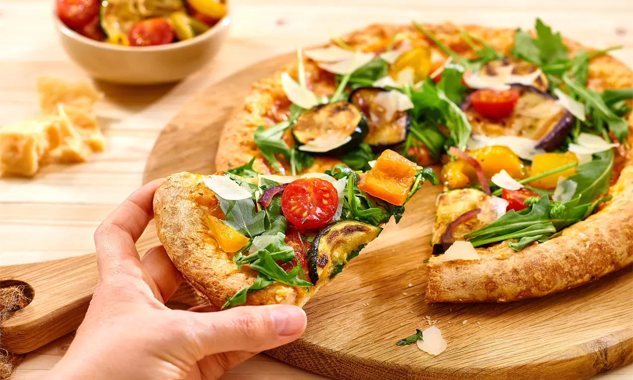 Goldbraune Pizza mit knusprigem Rand, belegt mit farbigem Gemüse, frischem Rucola und Parmesansplittern auf einem Holzbrett
