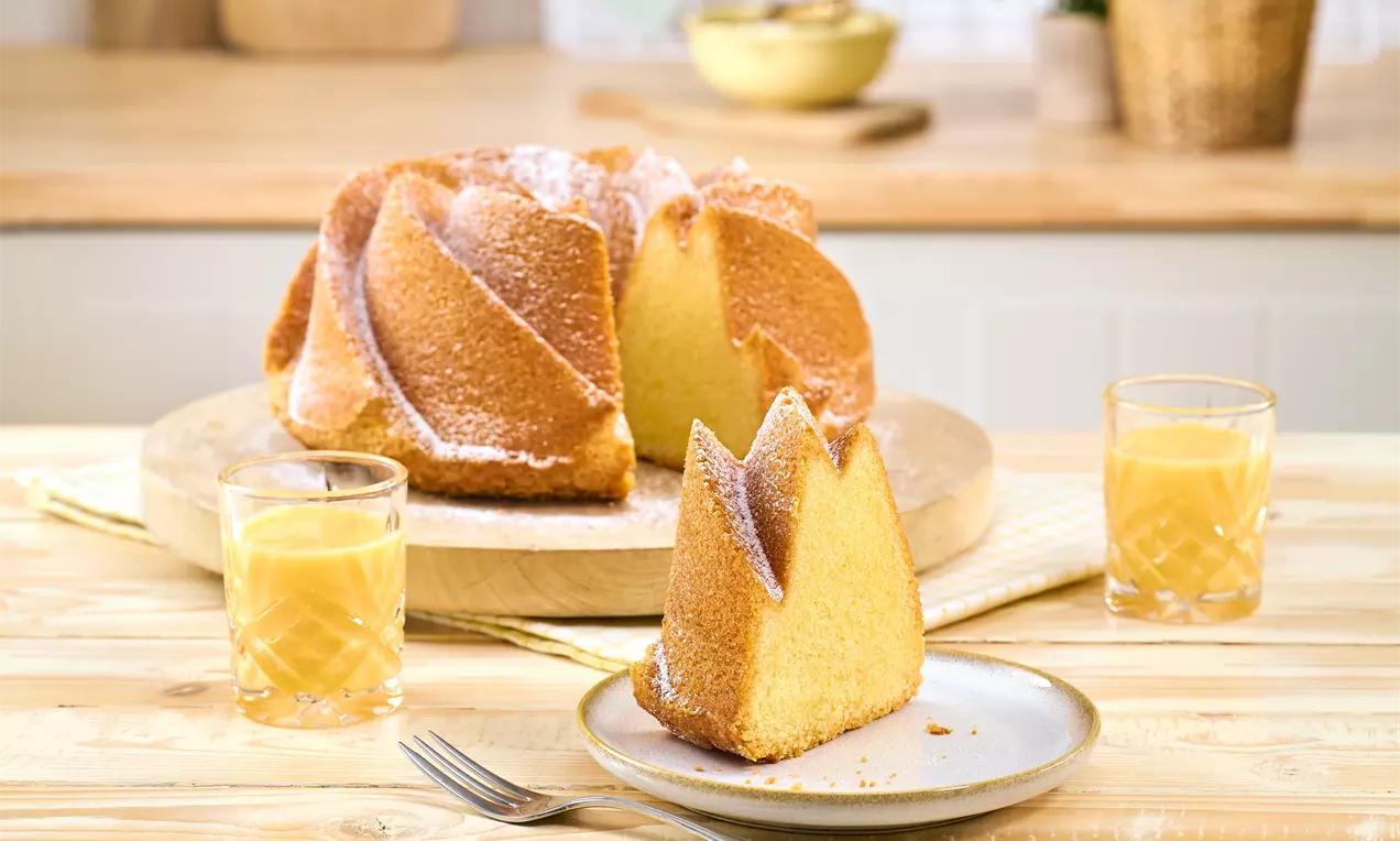 Eierlikörkuchen aus einer Twist-Gugelhupfform, goldbraun gebacken und mit Puderzucker bestäubt. Ein angeschnittenes Stück steht auf einem Teller, daneben zwei Gläser Eierlikör.