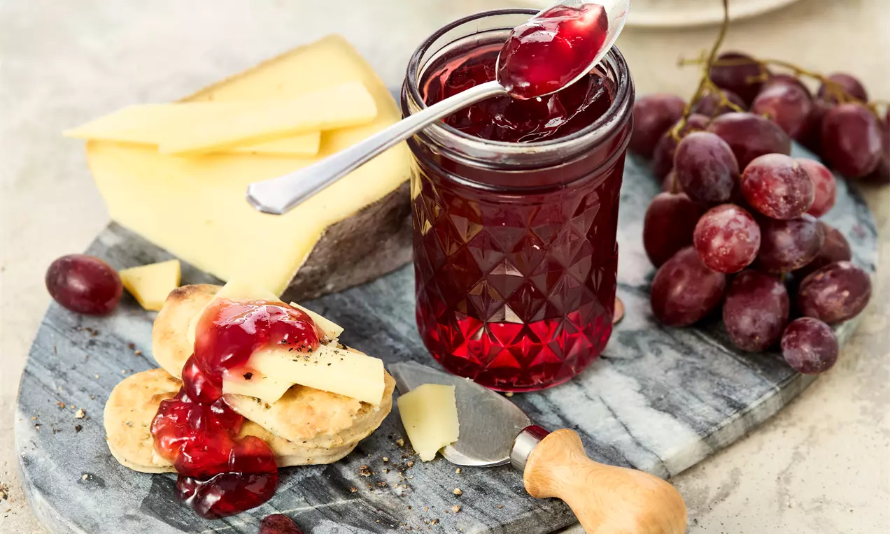 Glas mit rotem Traubengelee auf einem Brett, daneben frische rote Trauben, Käse und Cracker. Eine Hand gibt Gelee mit einem Löffel auf Cracker. Im Hintergrund ein weiteres Glas und Teller.