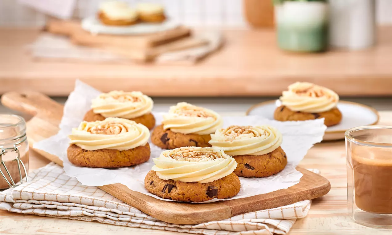 Runde braune Cookies mit dunklen Schokostückchen, gekrönt von cremig beigem Topping mit Zimtsprenkeln, serviert auf Holzbrett mit Papier