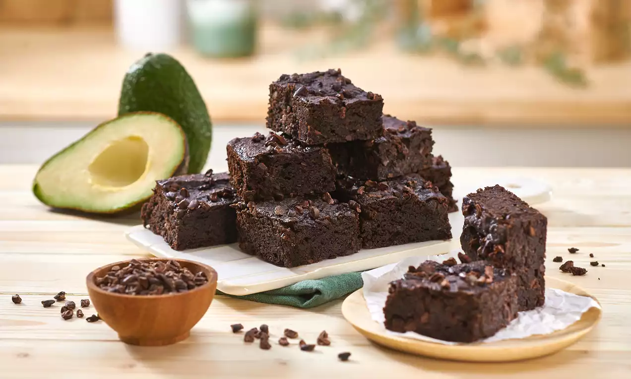 Saftige dunkelbraune Brownies mit glänzenden Kakaostückchen, gestapelt auf einem weißen Teller neben einer halbierten Avocado und einem Holzschälchen