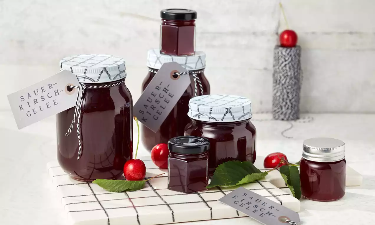Mehrere Gläser mit dunkelrotem Sauerkirsch-Gelee, teils mit karierten Deckeln und Etiketten, arrangiert auf einem weißen Untergrund mit Kirschen und Blättern
