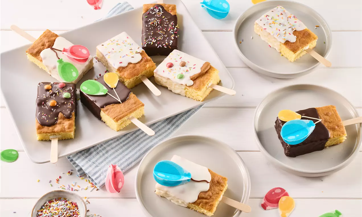 Kuchen am Stiel aus hellem Teig, verziert mit bunter Glasur, Streuseln und Deko in Luftballonform, auf Tellern und einer Platte angerichtet