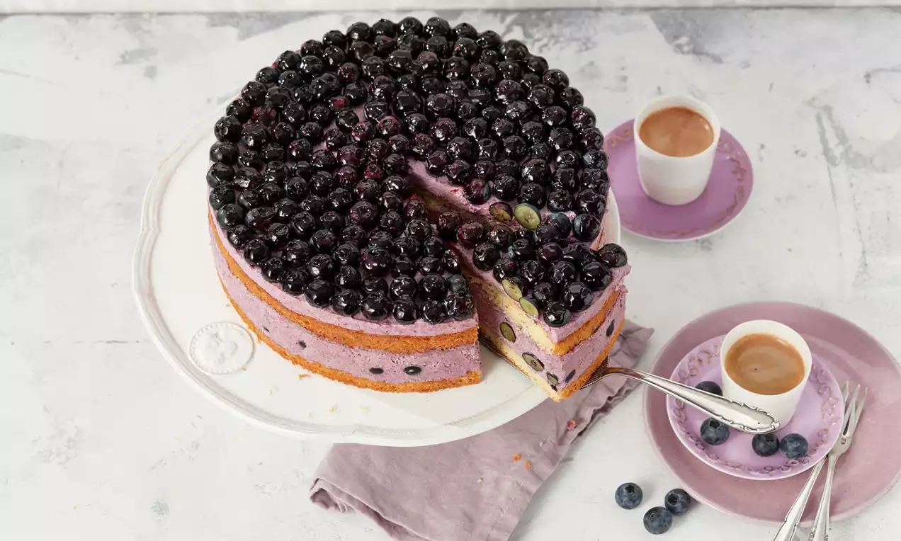 Blaubeertorte mit saftigem Biskuitboden, sahniger Joghurtcreme, glänzenden frischen Blaubeeren und grünem Minzblatt auf weißem Teller