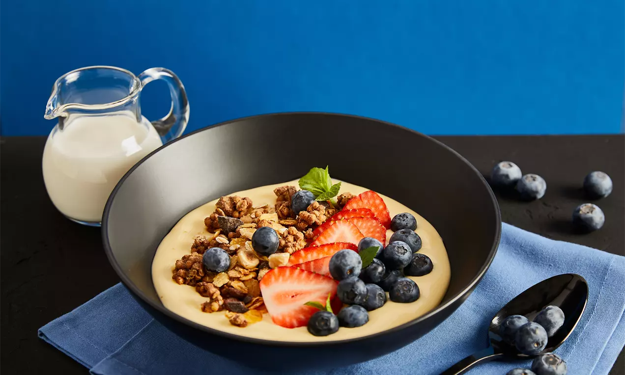 Protein-Müsli in einer schwarzen Schale mit cremiger gelber Basis, frischen Erdbeerscheiben, Blaubeeren, Müslistreuseln und einem Minzblatt