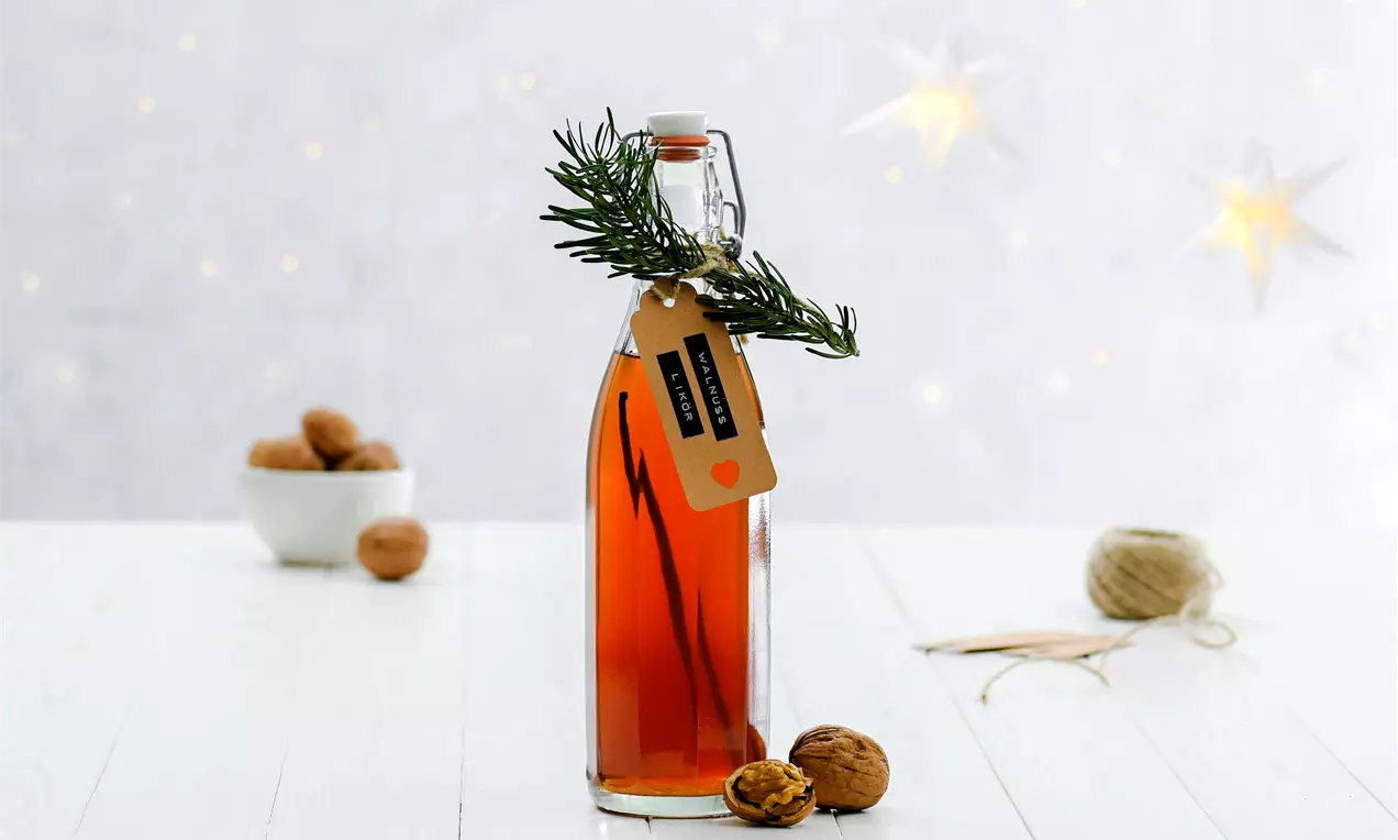 Eine schlanke Glasflasche mit goldbraunem Walnusslikör und Papieretikett, verziert mit grünem Tannenzweig, umgeben von Walnüssen auf hellem Holz