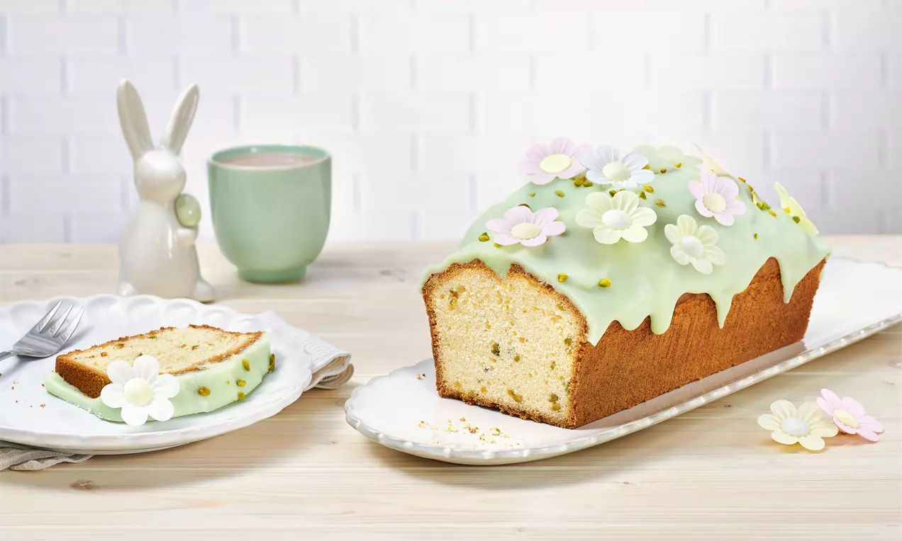 Saftiger Kastenkuchen mit hellem Teig, zartgrünem Zuckerguss, bunten Blüten aus Zucker und Pistazienstreuseln auf einer weißen Platte