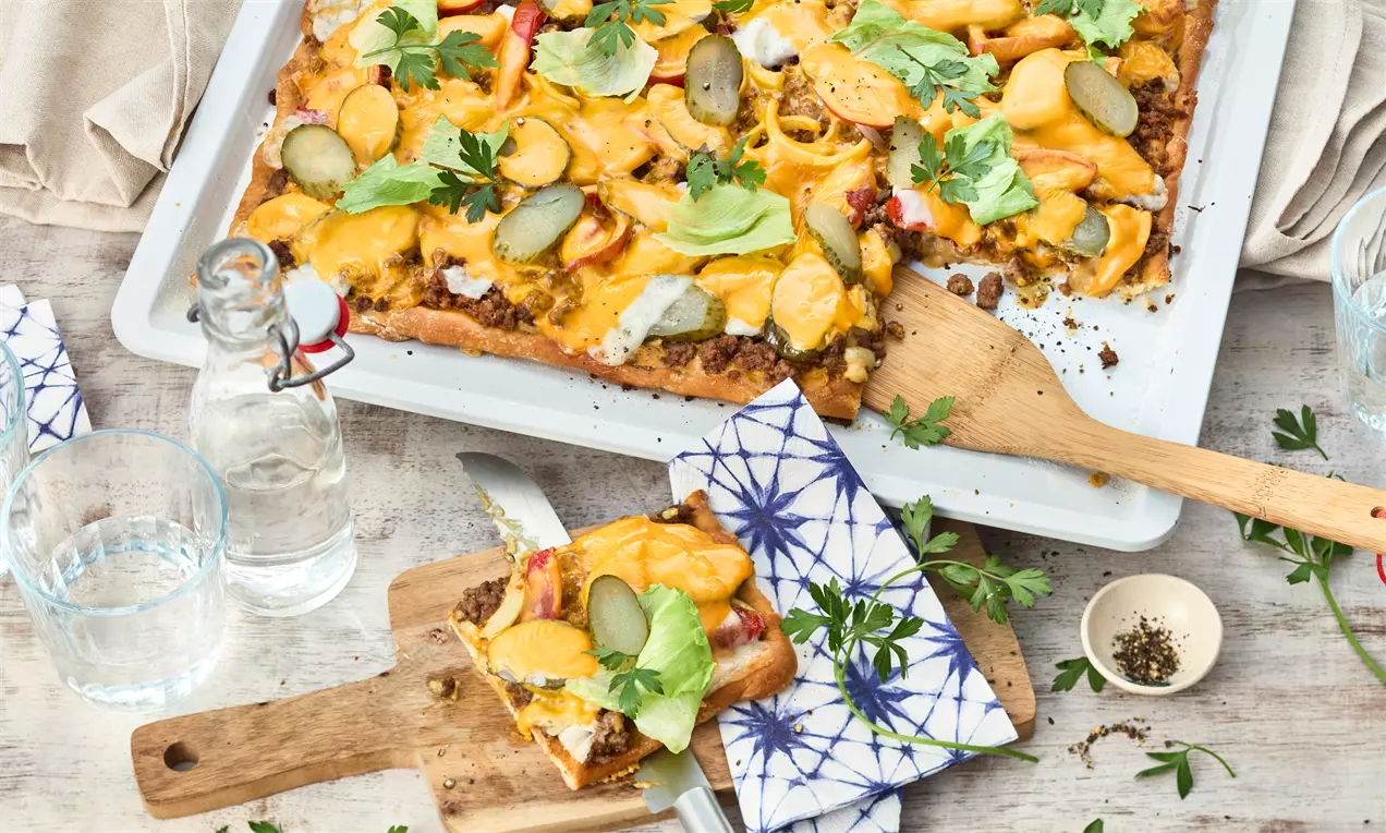 Rechteckige Pizza mit goldbraunem Käse, salatgrünen Blättern, Gurkenscheiben und Hackfleisch auf einem Blech und Holzbrett angerichtet