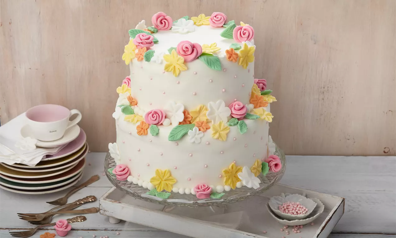 Weiße zweistöckige Hochzeitstorte, verziert mit rosa Rosen, gelben und weißen Blüten sowie grünen Blättern auf Glasplatte und Holzuntergrund