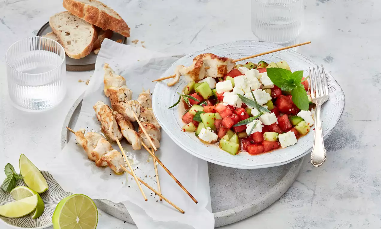 Frischer Melonen-Feta-Salat mit grünen Gurkenwürfeln, weißen Feta-Stücken und Minzblättern auf weißem Teller neben gegrillten Hähnchenspießen