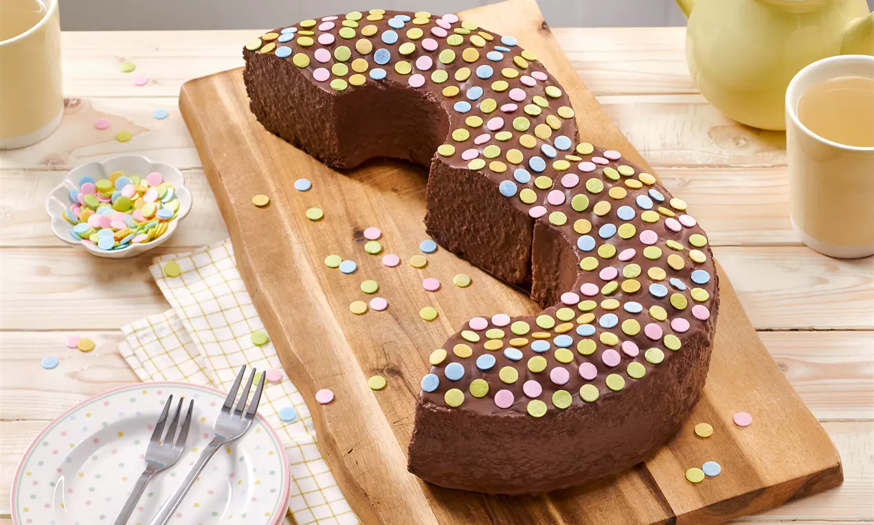 Schokoladenkuchen in Form einer Drei mit bunter Zuckerstreusel-Dekoration auf einem Holzbrett, daneben Teller und Teetasse